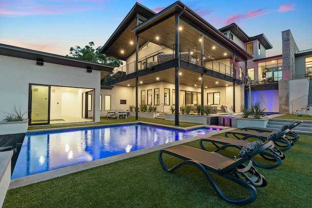 back of house at dusk with a patio, a yard, an outdoor pool, and a balcony