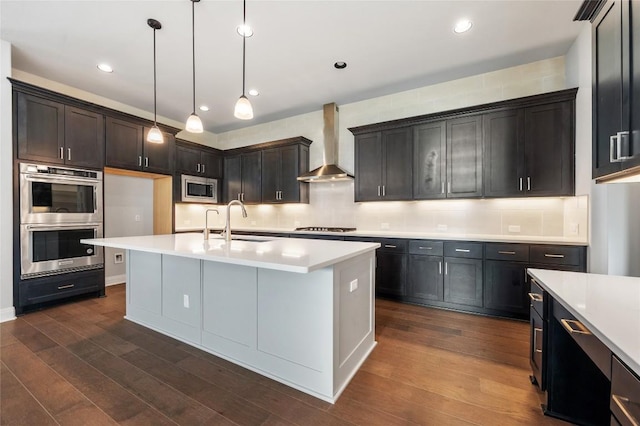 kitchen with a sink, wall chimney range hood, dark wood finished floors, appliances with stainless steel finishes, and light countertops