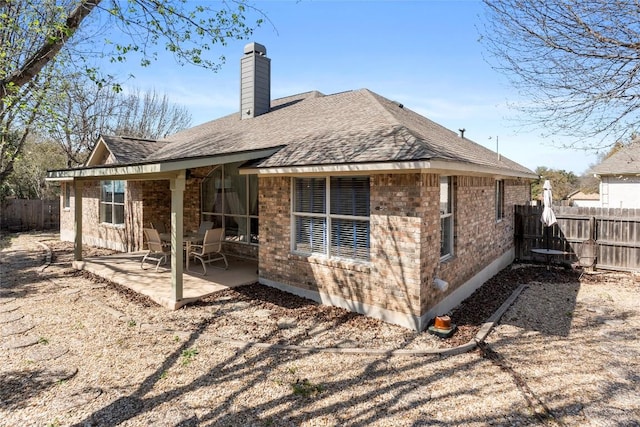 rear view of house featuring a patio, fence, roof with shingles, brick siding, and a chimney