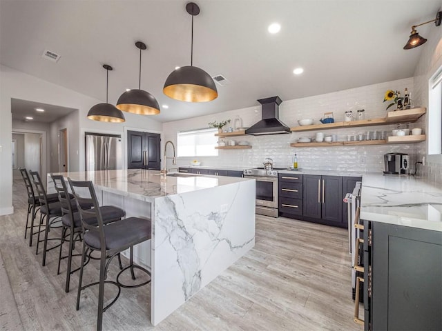 kitchen with open shelves, wall chimney range hood, appliances with stainless steel finishes, a sink, and light stone countertops
