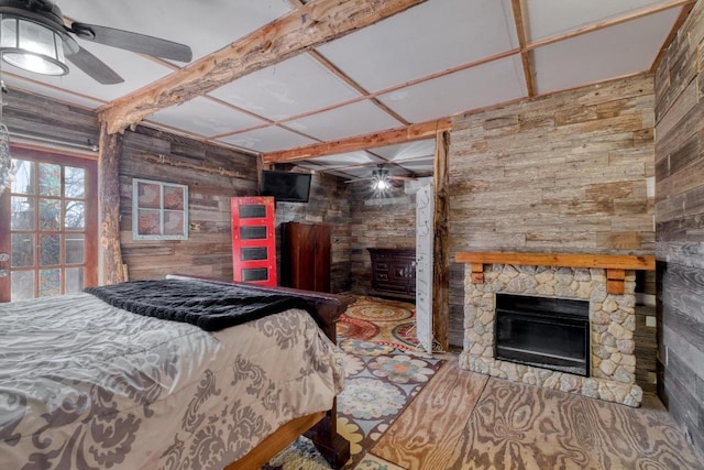 bedroom featuring beam ceiling, wood finished floors, a stone fireplace, wood walls, and ceiling fan