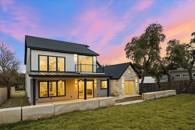 back of house at dusk with board and batten siding, a fenced backyard, a balcony, a yard, and a standing seam roof