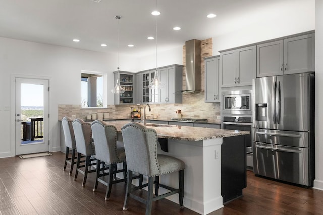 kitchen with light stone counters, gray cabinets, appliances with stainless steel finishes, wall chimney exhaust hood, and a kitchen island with sink