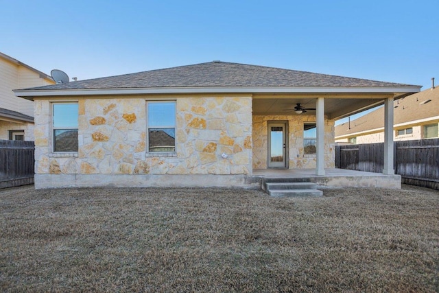 rear view of property with stone siding, a shingled roof, a ceiling fan, and fence