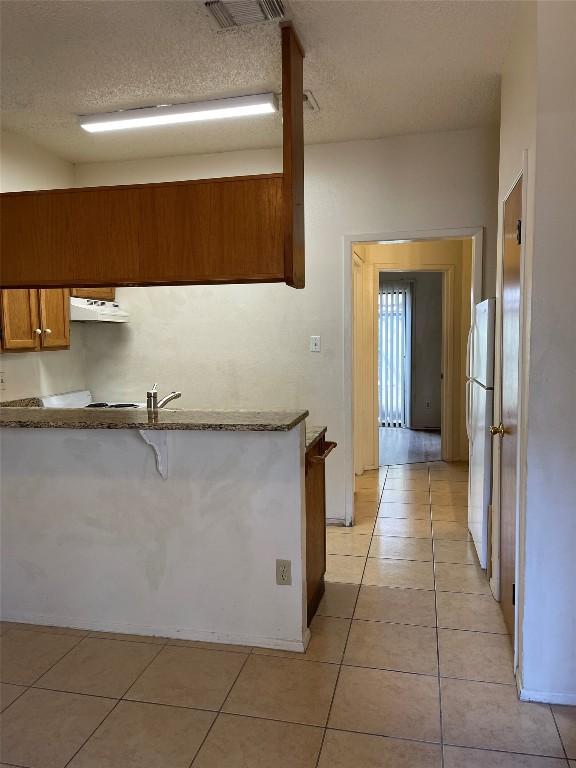 kitchen with under cabinet range hood, brown cabinets, light tile patterned floors, and freestanding refrigerator