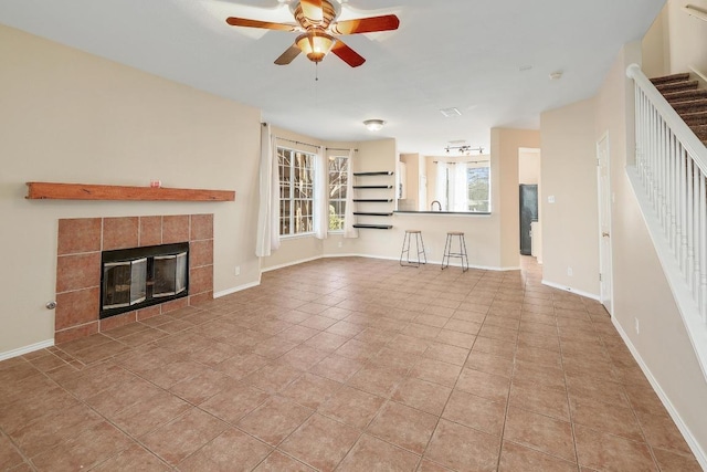 unfurnished living room with tile patterned floors, a ceiling fan, a tiled fireplace, baseboards, and stairs