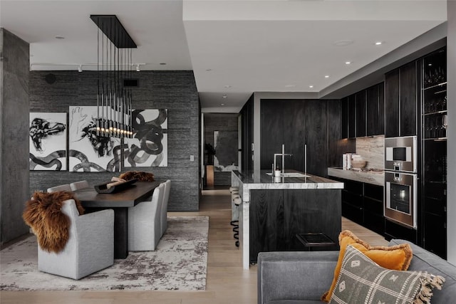 kitchen featuring light wood finished floors, a center island with sink, stainless steel oven, dark cabinetry, and modern cabinets