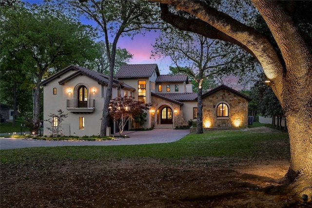 mediterranean / spanish home featuring a tiled roof, stucco siding, stone siding, a front lawn, and a balcony
