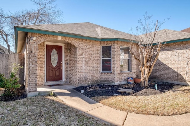 doorway to property featuring brick siding, roof with shingles, and fence