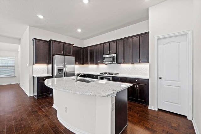 kitchen featuring light stone countertops, a center island with sink, dark wood-style flooring, a sink, and appliances with stainless steel finishes