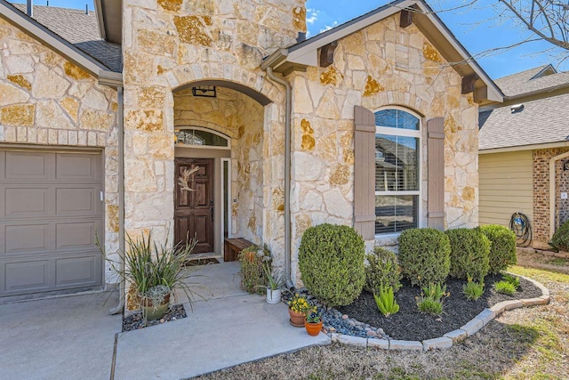 view of exterior entry with an attached garage, stone siding, and roof with shingles