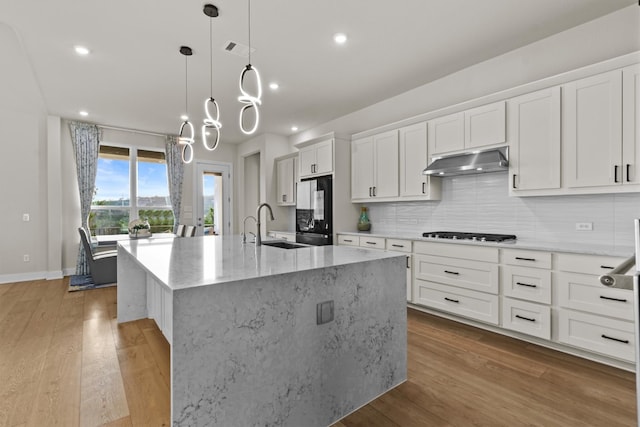 kitchen with a kitchen island with sink, a sink, light wood-style floors, under cabinet range hood, and tasteful backsplash