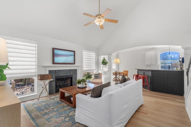 living room featuring light wood-style flooring, a tile fireplace, arched walkways, high vaulted ceiling, and ornate columns