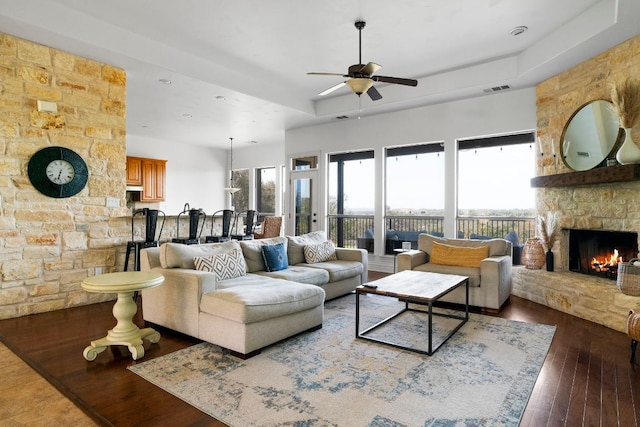 living room featuring a ceiling fan, visible vents, a tray ceiling, a stone fireplace, and hardwood / wood-style flooring