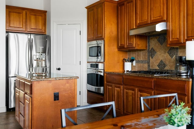 kitchen with under cabinet range hood, dark stone countertops, backsplash, a center island, and stainless steel appliances