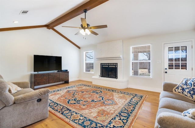 living area featuring visible vents, a brick fireplace, lofted ceiling with beams, light wood-style flooring, and a ceiling fan