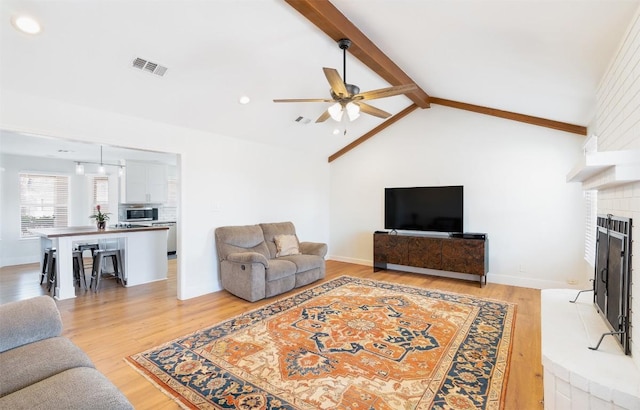 living room with visible vents, light wood-style flooring, lofted ceiling with beams, baseboards, and a brick fireplace