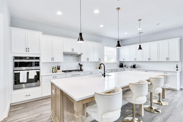 kitchen with tasteful backsplash, a sink, under cabinet range hood, double oven, and a kitchen island with sink