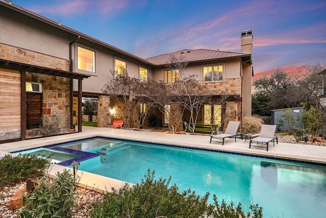 back of house at dusk featuring a patio, a pool with connected hot tub, a chimney, stucco siding, and stone siding