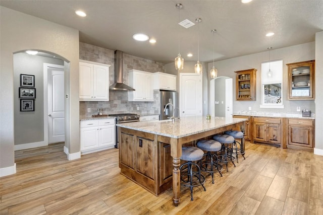 kitchen with a breakfast bar area, light wood-style flooring, arched walkways, stainless steel appliances, and wall chimney exhaust hood