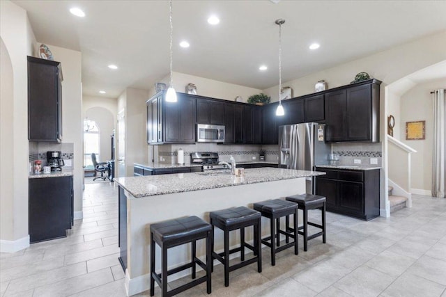 kitchen with light stone counters, a breakfast bar, an island with sink, arched walkways, and appliances with stainless steel finishes