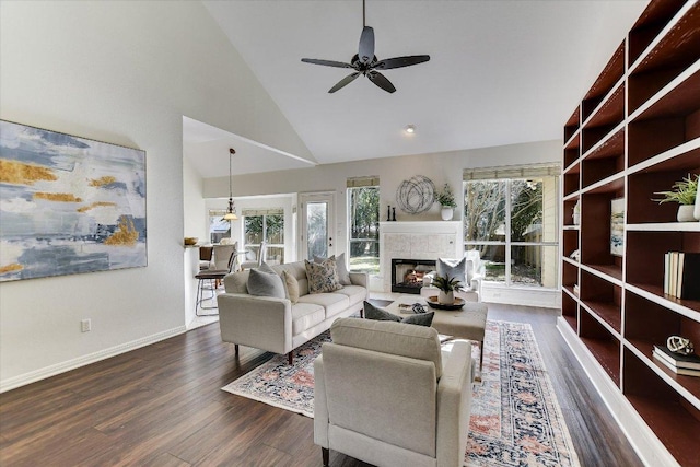 living room featuring a ceiling fan, baseboards, high vaulted ceiling, dark wood-style flooring, and a lit fireplace