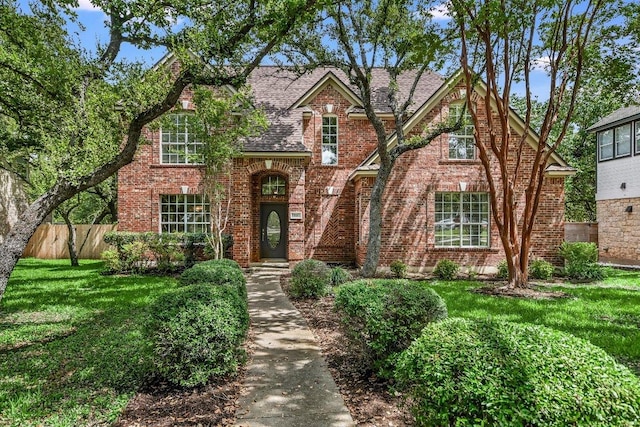 english style home featuring brick siding, a shingled roof, a front lawn, and fence