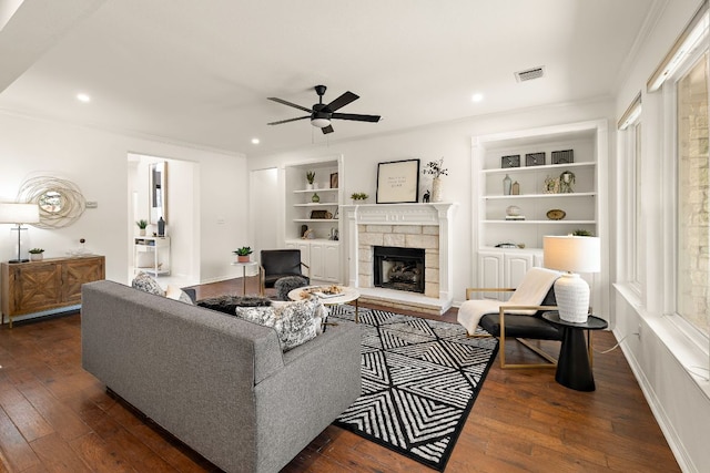 living room with built in shelves, visible vents, dark wood-style flooring, ceiling fan, and a stone fireplace