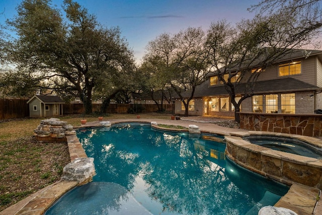 pool at dusk featuring an outbuilding, a shed, a pool with connected hot tub, and a fenced backyard