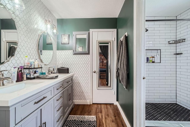 full bathroom featuring wood finished floors, tiled shower, wallpapered walls, double vanity, and a sink