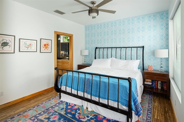 bedroom featuring wood finished floors, visible vents, baseboards, wallpapered walls, and an accent wall