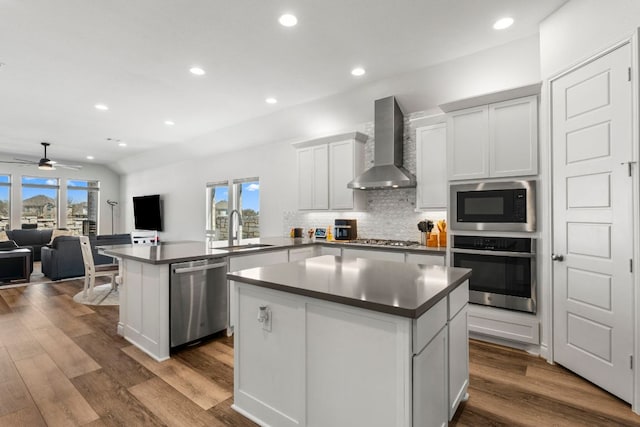 kitchen with a sink, a kitchen island, a peninsula, appliances with stainless steel finishes, and wall chimney exhaust hood