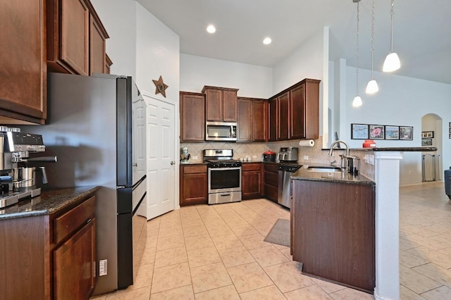 kitchen featuring a sink, stainless steel appliances, arched walkways, dark stone counters, and a peninsula