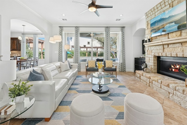 living room featuring ornamental molding, ceiling fan with notable chandelier, arched walkways, a stone fireplace, and light tile patterned floors