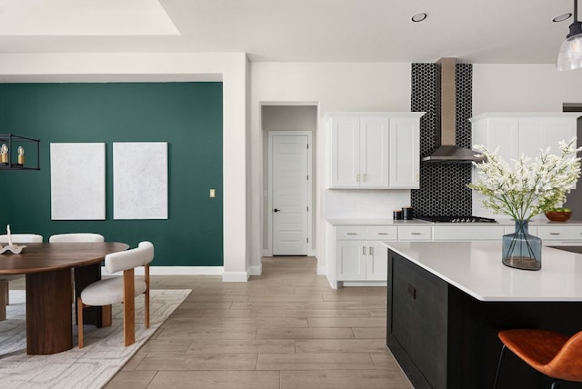 kitchen with wall chimney range hood, tasteful backsplash, white cabinets, light wood finished floors, and light countertops