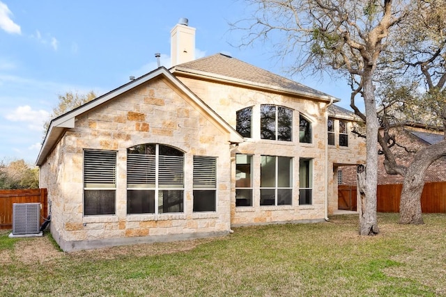 rear view of house featuring fence, central AC, a chimney, stone siding, and a lawn