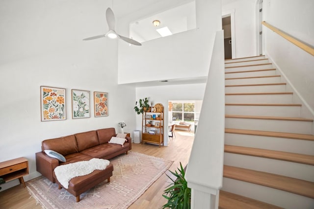 living room featuring stairs, light wood-style flooring, a skylight, a towering ceiling, and a ceiling fan