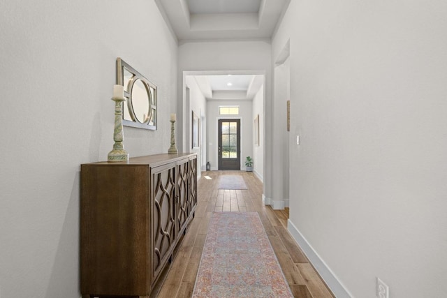 corridor with a tray ceiling, baseboards, and light wood finished floors