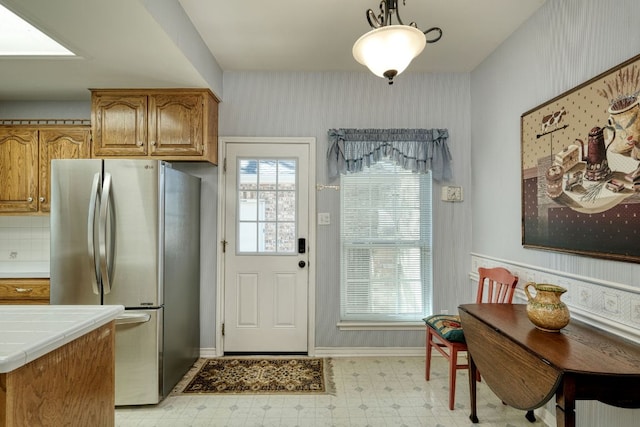 kitchen with brown cabinetry, light floors, wallpapered walls, freestanding refrigerator, and tile counters