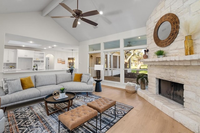 living room with wood finished floors, a ceiling fan, beam ceiling, arched walkways, and a stone fireplace