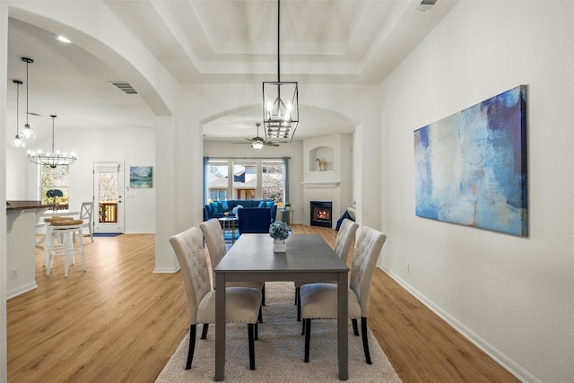 dining area with visible vents, a warm lit fireplace, light wood-style flooring, and ceiling fan with notable chandelier