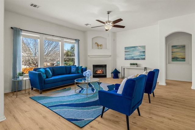 living room with visible vents, baseboards, a warm lit fireplace, wood finished floors, and a ceiling fan