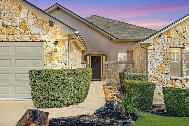 doorway to property featuring a garage, stone siding, roof with shingles, and stucco siding