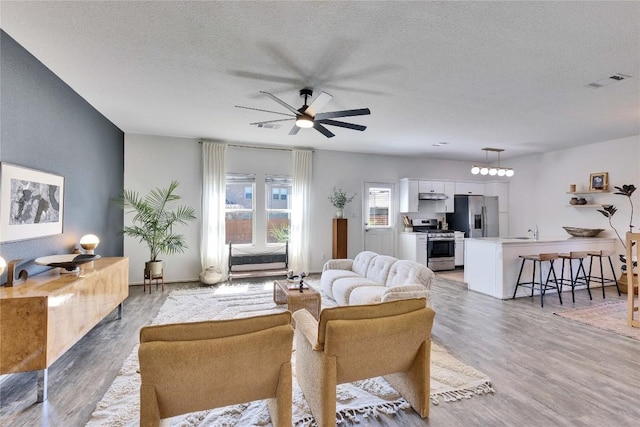 living room featuring a ceiling fan, visible vents, light wood finished floors, and a textured ceiling