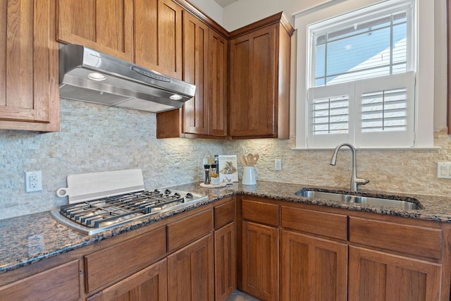 3129 Mill Stream Dr, Cedar Park, TX 78613 - kitchen with under cabinet range hood, brown cabinetry, stainless steel gas stovetop, backsplash, and dark stone countertops