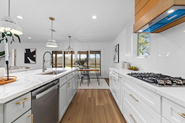 kitchen featuring wood finished floors, a sink, stainless steel appliances, wall chimney range hood, and decorative light fixtures