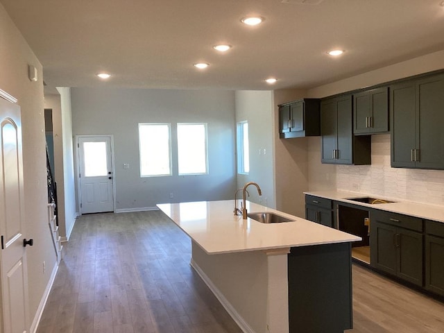 kitchen with black electric stovetop, a center island with sink, decorative backsplash, light wood-style flooring, and a sink