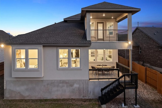 rear view of house featuring stucco siding, a balcony, roof with shingles, and fence