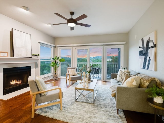 living room featuring wood finished floors, a warm lit fireplace, and ceiling fan