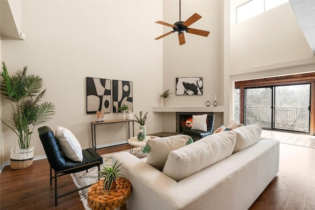 living area featuring baseboards, ceiling fan, a lit fireplace, a high ceiling, and wood finished floors